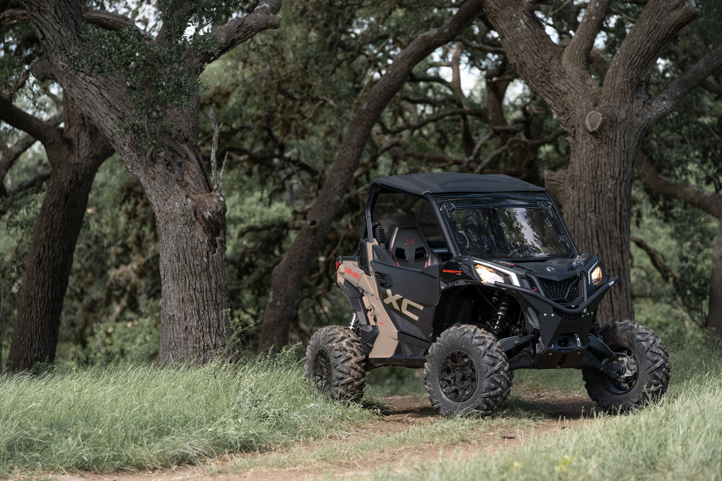 A Desert Tan and Carbon Black Can-Am Maverick Sport X XC parked in a forest setting 